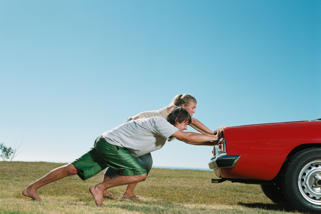 Homem e mulher empurram traseira de carro vermelho num prado verde